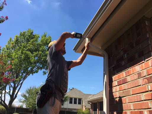 rain gutters are being installed. we can see gutter technician attaching a downspout for water drainage for the rain gutter system. blue sky and brick wall house. green bushes on the background. 